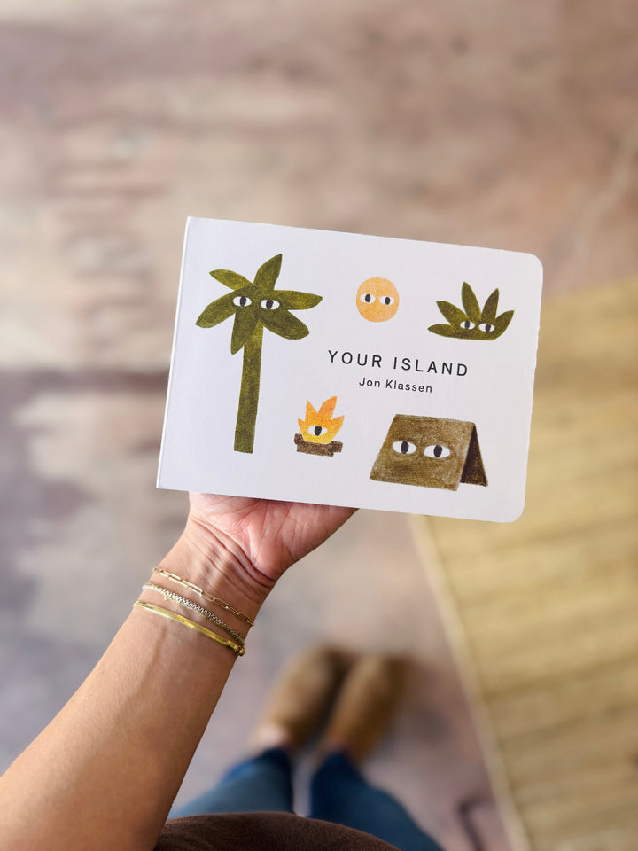 Hand holding a Your island by jon klassen with illustrations of a palm tree, sun, and beach bag